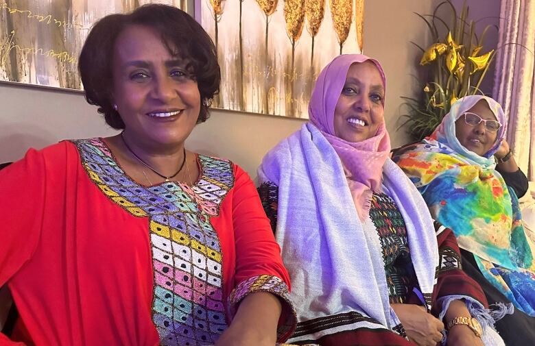 Three women wearing colourful dresses sit together on a couch. 