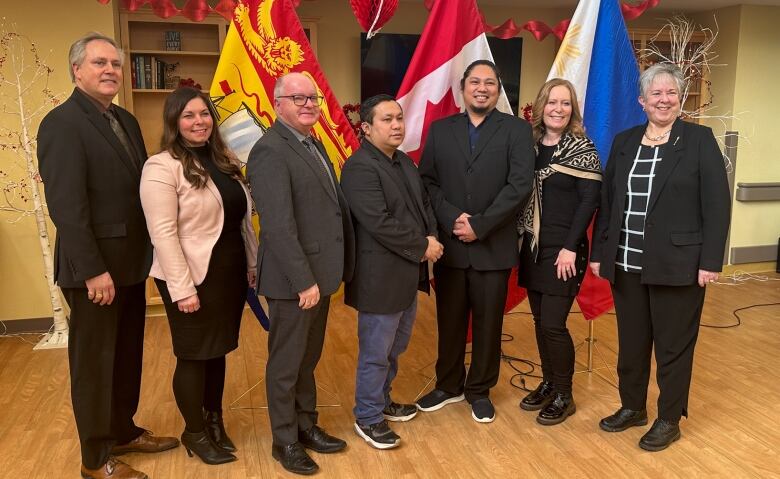 Seven people stand side by side in front of a three flags. 