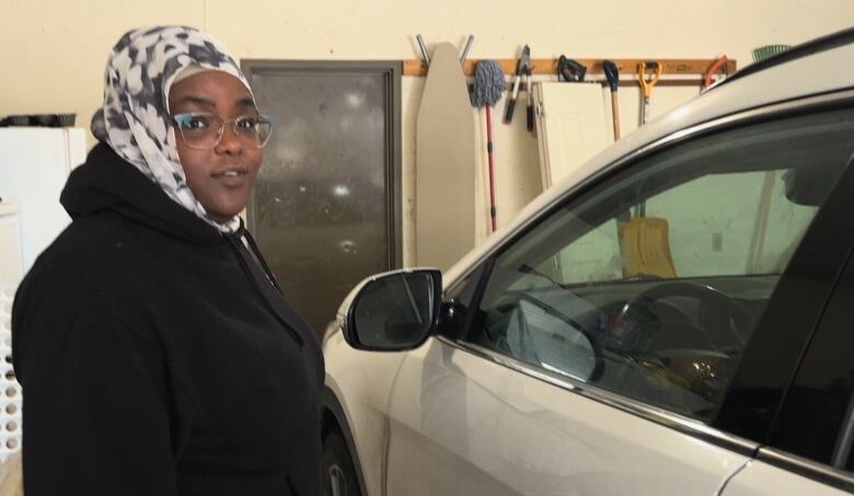 A woman wearing a hijab stands beside a vehicle in a garage.