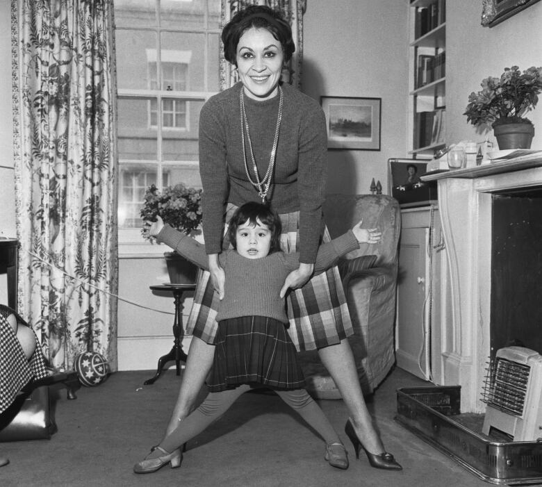 A smiling woman in a living room stands in front of a small child, holding her under her armpits. The photo is in black and white.