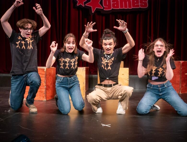 Four students perform on stage with scared looks on their faces, with their hands raised. 