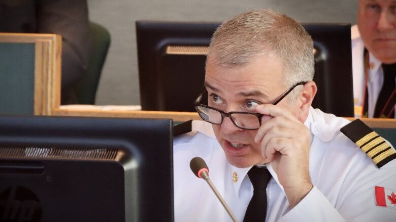 A man in a white uniform with black and gold shoulder patches pulls his dark-framed glasses down and looks over them in front of a microphone.