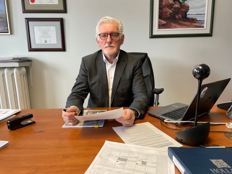 Man with white hair and beard wears glasses and looks at the camera while sitting at a desk.