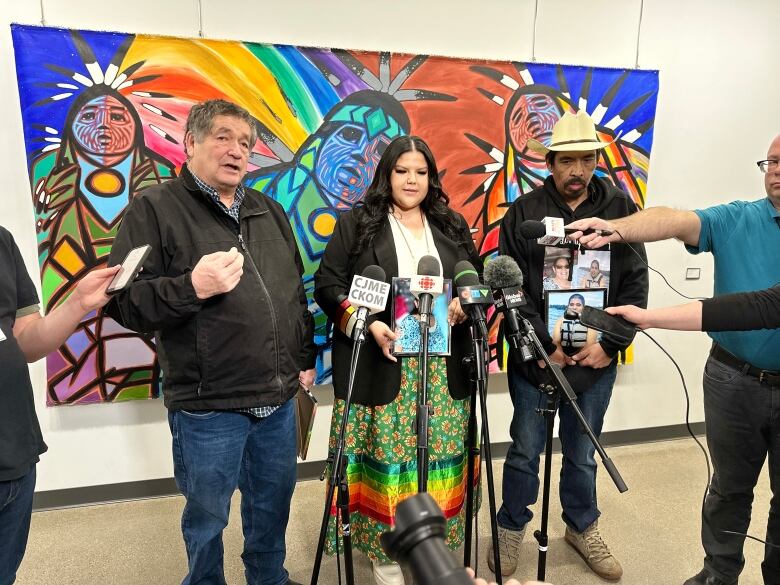 Two men flank a woman at a collection of microphones during a media conference.