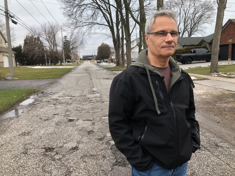 A man with grey hair wearing glasses and a black jacket standing on an old, cracked road