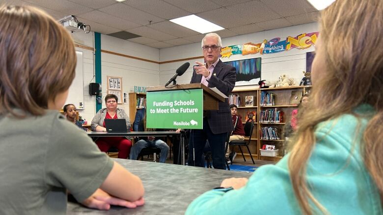 Children sit at tables, looking at a man behind a podium that says 'Funding schools to meet the future.' There are bookshelves behind him.