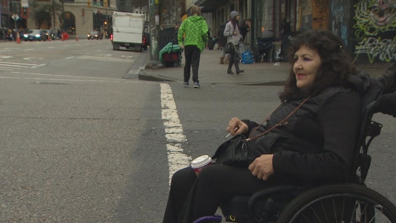 A person smokes a cigarette with a coffee between her knees on a downtown street.