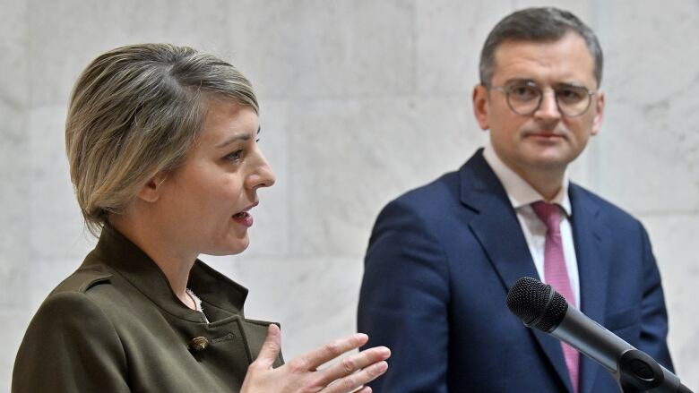 A woman in a brown suit speaks into a microphone while a man in a blue suit watches from the side. 