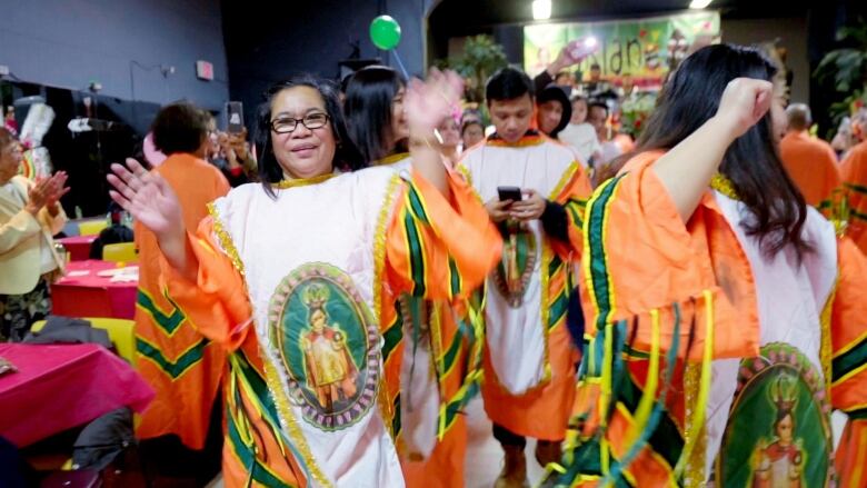 Group of pwoplw wearing bright orange and white smocks dance in community centre. Woman in front has long dark hair and smiles with her hands up, palms forward.