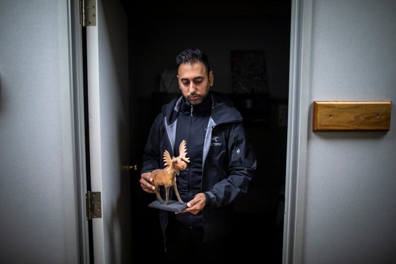 A South Asian man stands in a doorway holding a small wooden carving of a moose.