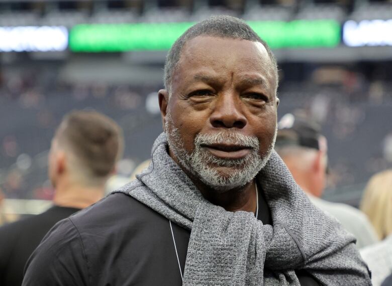 A man with a grey sweater folded around his neck smiles slightly at the camera. 