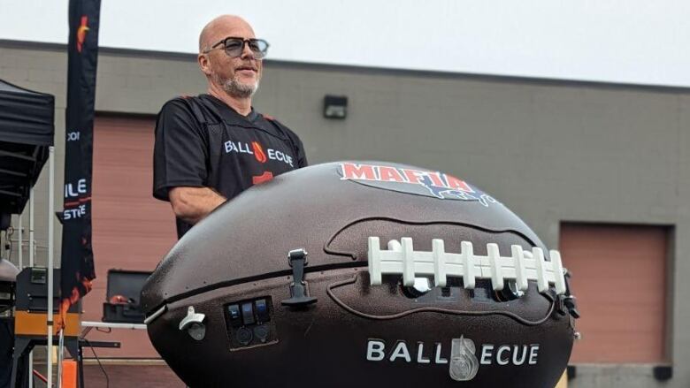 A man wearing a sports shirt, rolls a barbecue in the shape of a football. It says ballbecue on the side. 