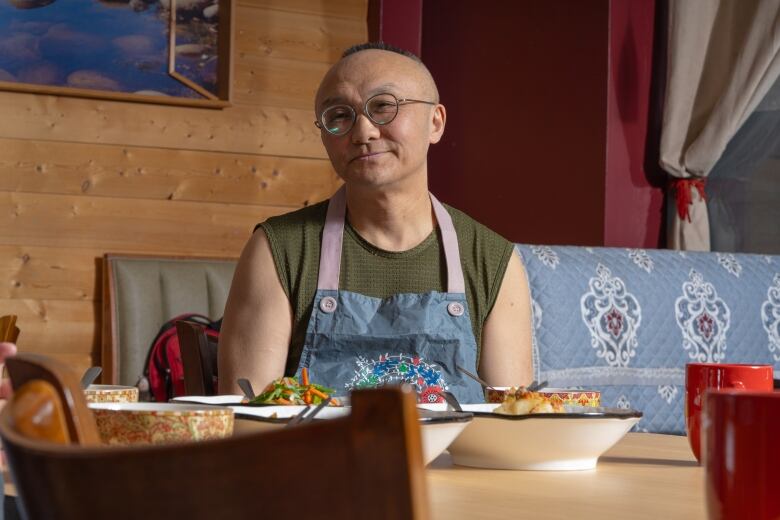 A Chinese man in an apron sits at a table in a restaurant with filled dishes displayed before him.