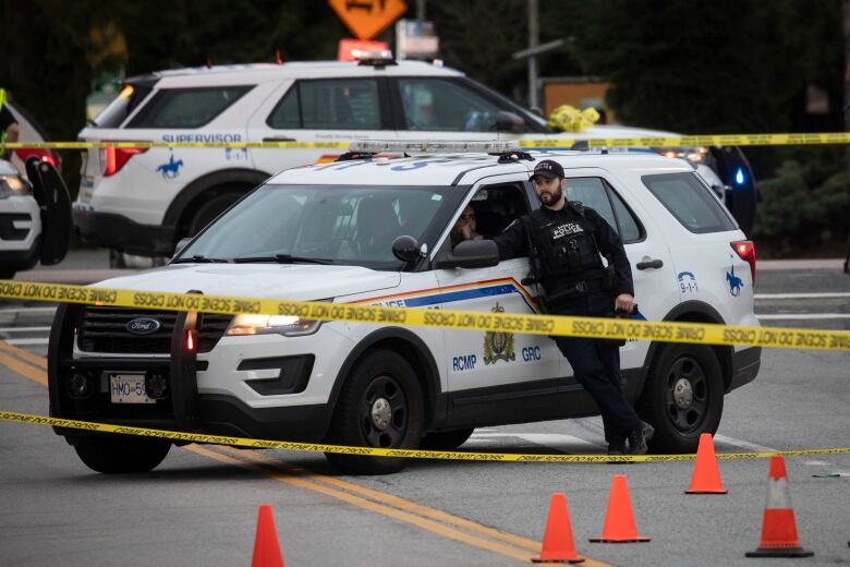 A police officer leans on a white police car, behind yellow crime tape.