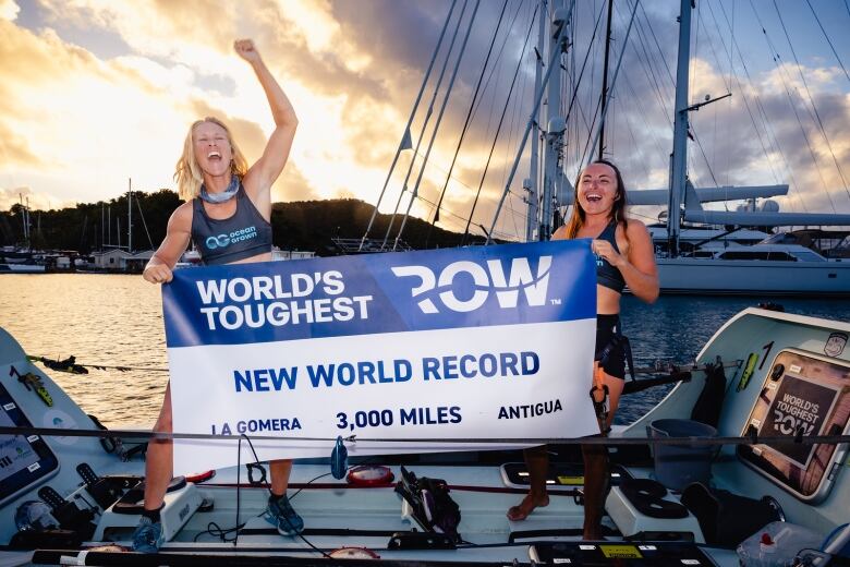 Two women in swimsuits on a boat deck hold up a banner that says New World Record.