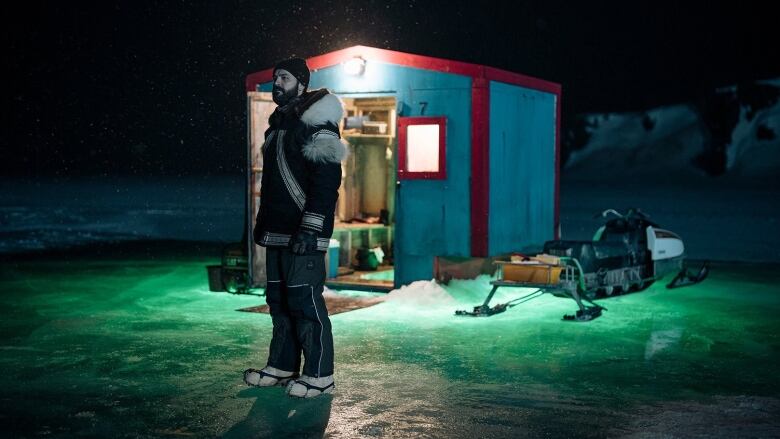 A man wearing a fur jacket is standing on top of ice in front of a smack shack, with the northern lights in the background.