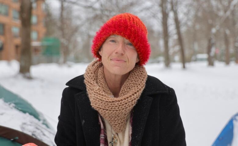 A woman wearing a red toque, knit scarf and black wool jacket looks straight into the camera with a snowy park behind her.