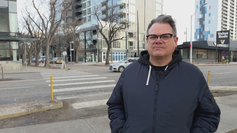 A white man with gray hair and black-rimmed glasses wearing a navy jacket standing on a sidewalk outside on a gloomy day. 