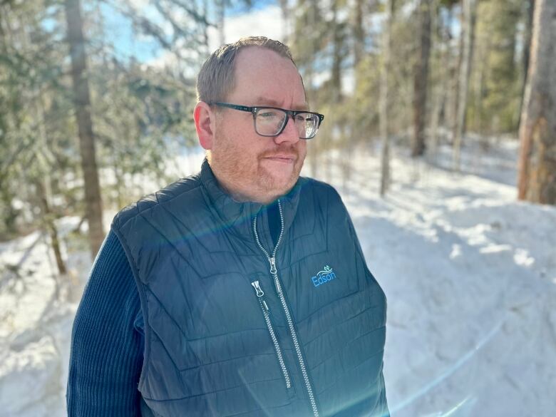 A man, with a blue coat and glasses, stands in the snow against some trees.