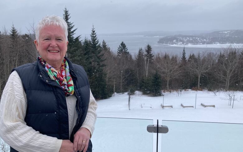 Woman with short white hair wearing quilted vest and sweater stands on a deck overlooking forest and ocean bay.