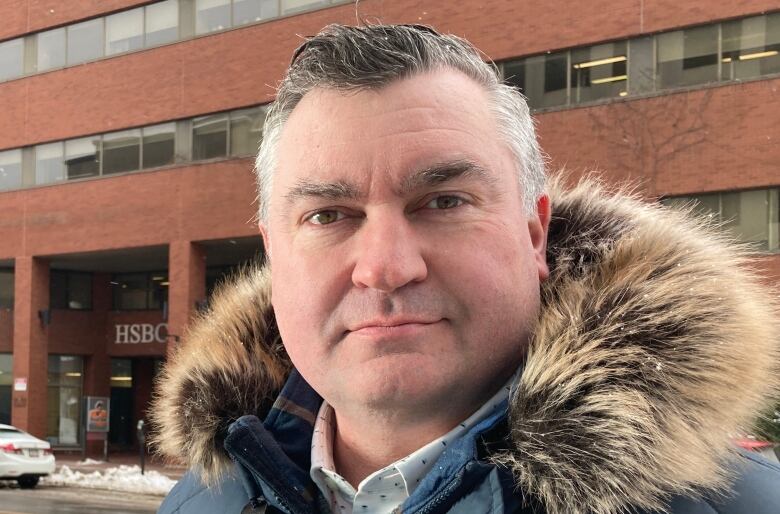 Man with short grey-brown hair and wearing winter parka stands in front of large downtown building.