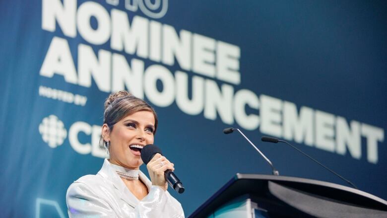 A smiling woman standing in front of a podium speaks into a microphone. Behind her the words 'Juno Nominee Announcement' can be seen in large type on a wall. 