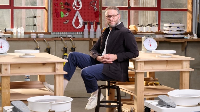 Seth Rogen sits on a stool between two wooden tables in a workshop. He's wearing a black jacket, glasses and jeans.