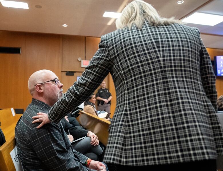 A bald man wearing glasses and a plaid shirt sits in a wood-paneled room and looks up at a blond woman in a plaid blazer who reaches out to touch his shoulder as an armed and uniformed deputy stands by the door. 