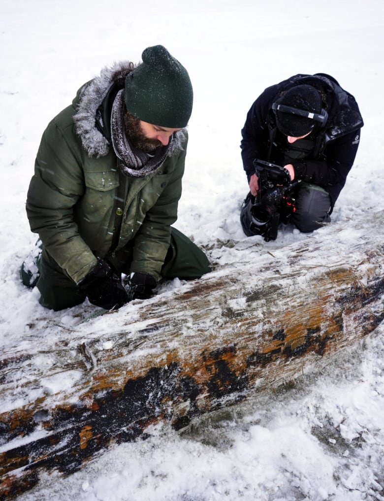 Two men out in snow, crouched down next to plank of wood.