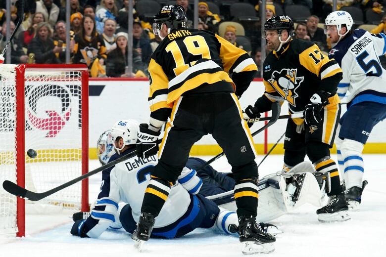 A Pittsburgh Penguin in black watches as a puck enters the net behind the Winnipeg Jets goalie and another player, both sitting on the ice.