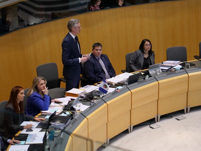 Man in suit stands in legislative assembly.