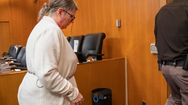 A woman wearing a white sweater and glasses walks toward out of a courtroom in handcuffs attached to a chain around her waist.