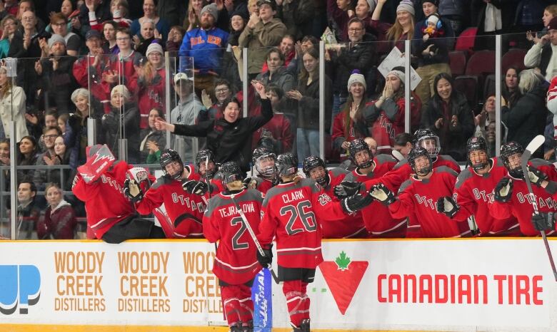 Two players in red hockey jerseys celebrate with their teammates on the bench. A crowd is seen behind them.