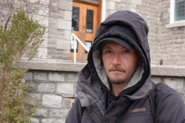 A man wearing a black hooded jacket and cap stares standing in front of a large, stone church building.