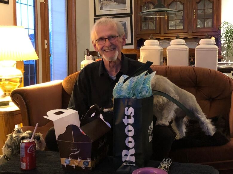 A man in a dark shirt and short grey hair smiles while sitting on a couch.