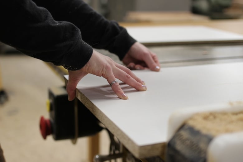 A woman's hand wearing an engineering ring guides a piece of wood through a saw.