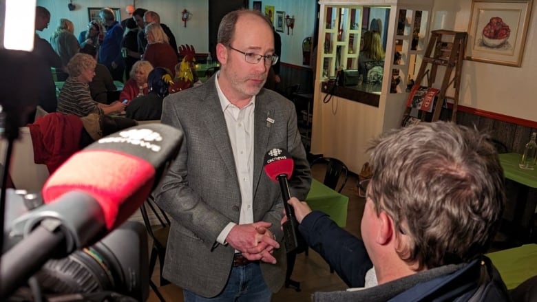 Balding man in glasses with beard and mustache talks into CBC microphone as Green Party workers celebrate behind him. 