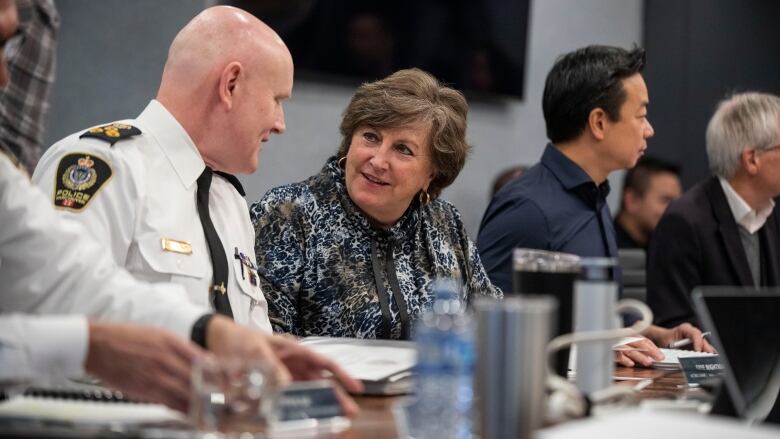 A bald man speaks with a white woman at a table, who is sitting next to an East Asian man.