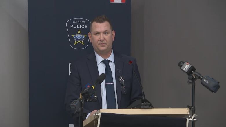 A white middle aged man in a suit stands in front of a police podium and microphone