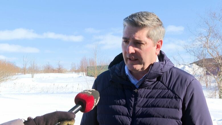 A man with short gray hair and a puffy winter jacket speaks into a microphone in and outdoor setting