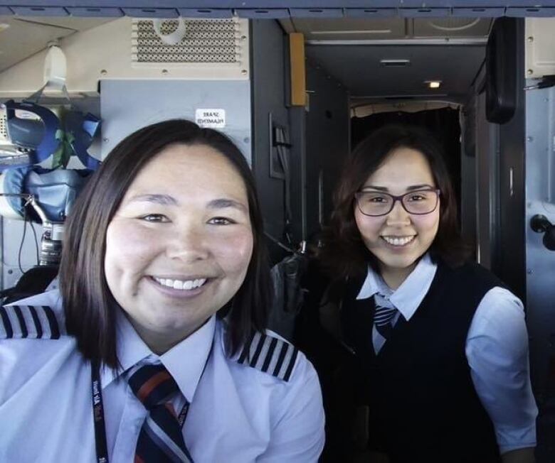 Two women take a selfie in the cockpit of an aircraft. One of them is in a flight attendant uniform, the other is in a pilot uniform.