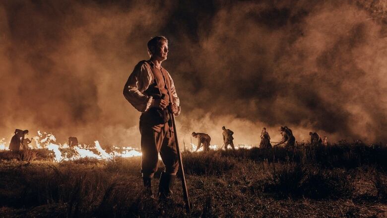 A man in historical garb stands in a field at night. Behind him people work the field. The field is partially ablaze.