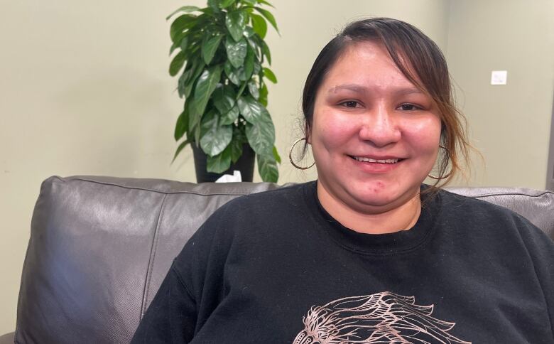 A woman with bangs, wearing a sweater, smiles at the camera. She's sitting on a leather armchair and there's a tall plant in the background.