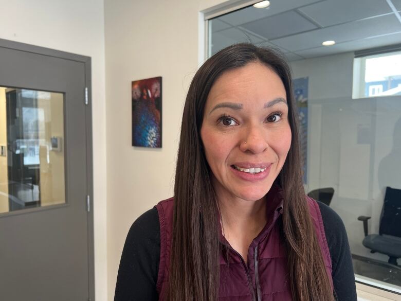 A woman with long hair smiles at the camera. She's in the entrance of a building. There's colourful art hanging on the wall behind her.