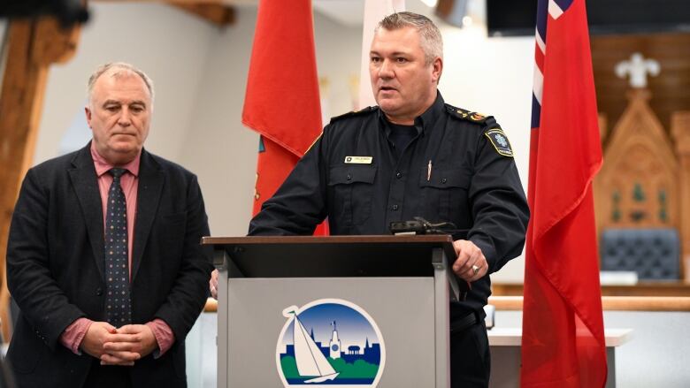 A man with short grey hair, waring a blue paramedic uniform stands at a podium. Behind him are The Canadian and Ontario flags and a large room with a council table and chairs.