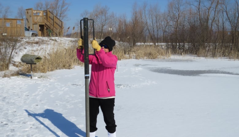 A person in winter clothes jams a pole into icy ground in a pond. 