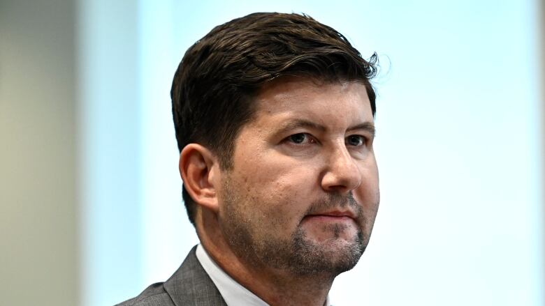 A man in a grey suit sits in front of a window.