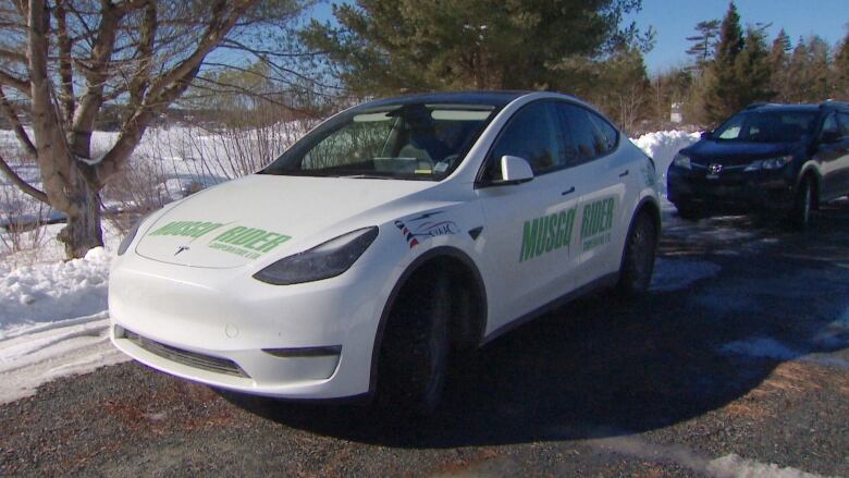 A white car sits in a gravel driveway with trees behind it