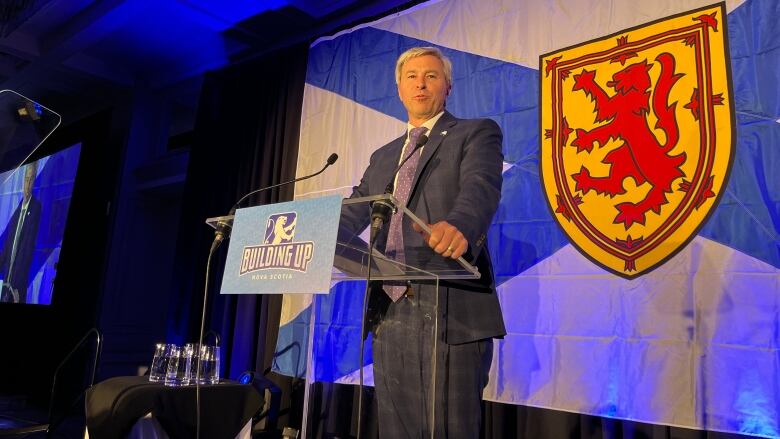 A man in a suit and tie stands at a podium with the Nova Scotia flag behind him.