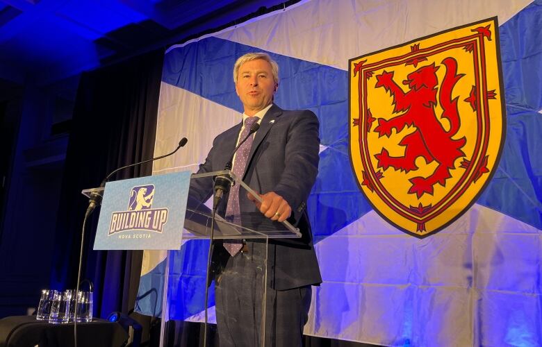 A man in a suit and tie stands at a podium with the Nova Scotia flag behind him.